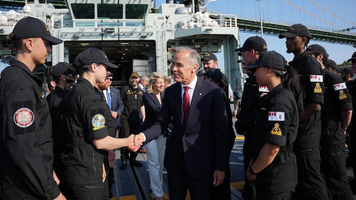 Mark Carney, sur un navire, est entouré d'hommes et de femmes en uniforme. Il serre la main d'un membre de la Marine.