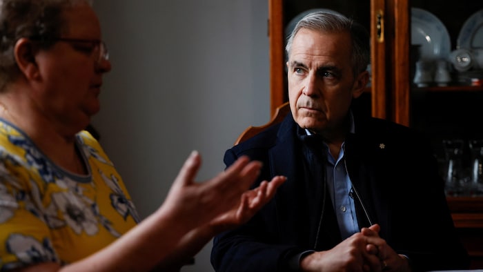 Mark Carney est assis à une table et écoute une citoyenne parler.