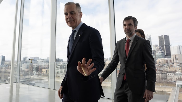 Mark Carney et Steven Guilbeault marchent devant des fenêtres qui montrent le centre-ville de Montréal.