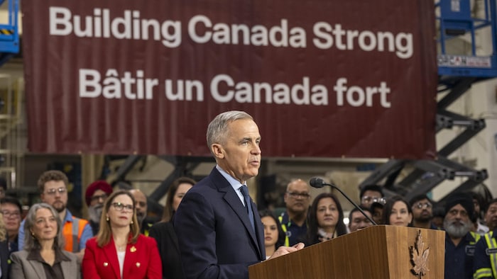 Mark Carney parle dans un micro lors d'une conférence de presse dans une usine.