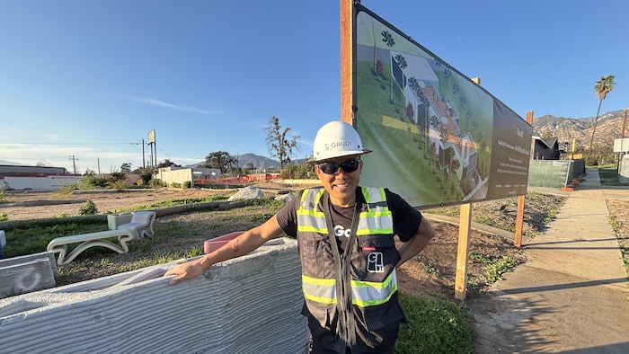 Un homme avec des habits de construction à côté d'un mur en béton imprimé et devant une affiche avec une maison modèle dans un quartier sinistré. 