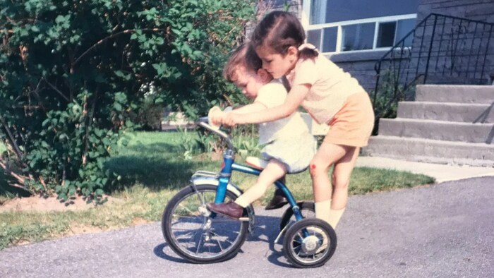 Un très jeune Mark Sutcliffe, sur un vélo, reçoit un coup de main de sa grande sœur.