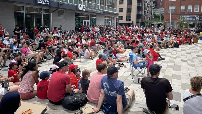 Une grande foule assise regarde vers un écran à la place Kassiwi.