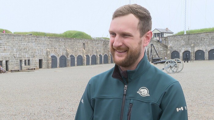 Un homme à la barbe rousse bien taillée est debout, entouré de fortifications en pierre, et sourit courtoisement.