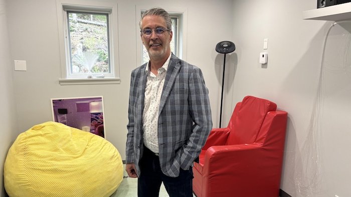 Martin Houde pose dans un petit salon. Des fauteuils aux couleurs vives ainsi qu'un projecteur s'y trouve.