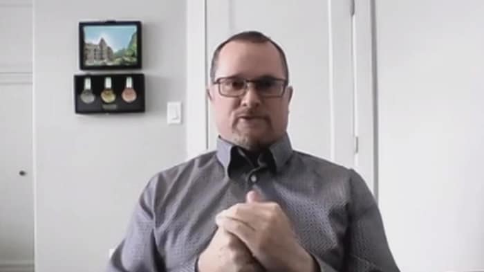 Un homme debout, les deux mains devant son corps, devant un mur  blanc.