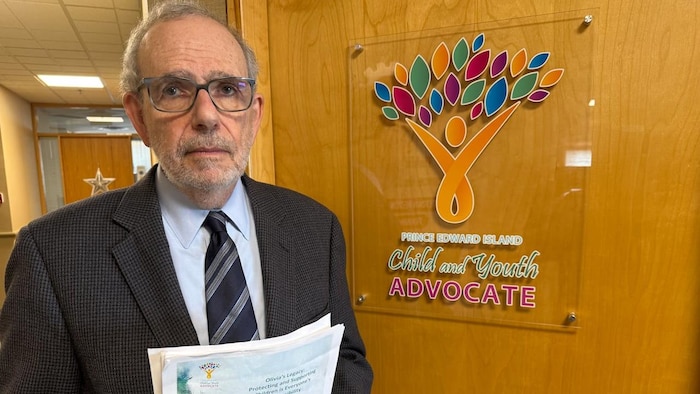 Marv Bernstein devant la porte de son bureau, tenant dans ses mains un rapport.
