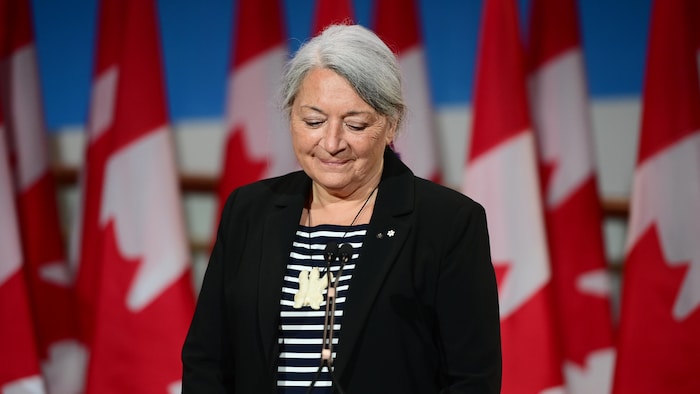 حاكمة كندا العامة ماري ماي سايمون واقفة وخلفها أعلام كندية.