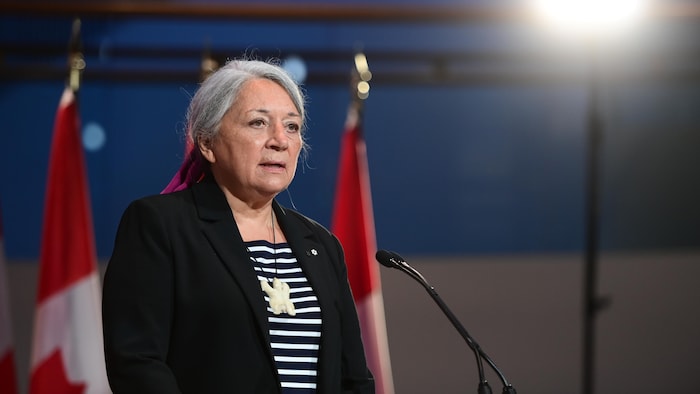 Mary Simon face à un micro, devant deux drapeaux canadiens.