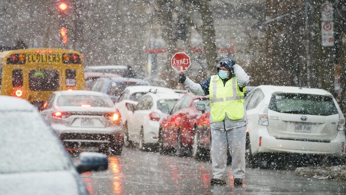 Une brigadière scolaire tient une pancarte d'arrêt sous une neige abondante dans une rue.