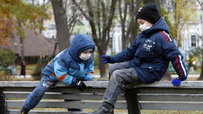 Deux enfants portant des masques grimpent sur le dossier d'un banc public dans un parc en automne.