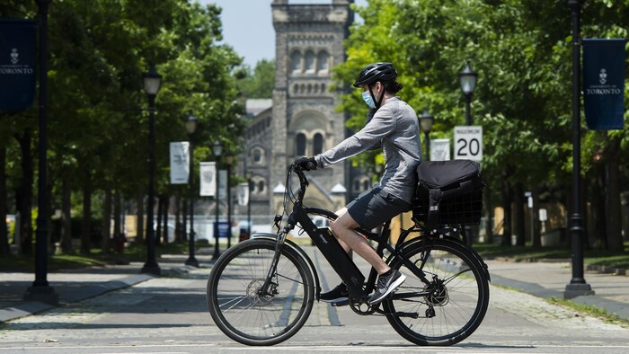 Un jeune qui porte un masque roule à vélo devant un bâtiment de l'Université de Toronto.