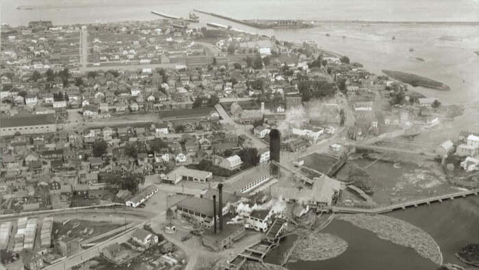 Photo en noir et blanc : on voit une partie du centre-ville incluant le quai qui donne accès au fleuve Saint-Laurent; en avant-scène, l'usine et sa longue cheminée fumante s'imposent dans le paysage.