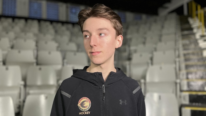 Un adolescent debout devant les gradins d'un aréna. Il regarde vers la gauche.