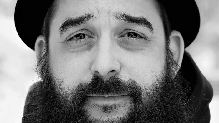Portrait en noir et blanc, extérieur, de l'auteur Mathieu Blais. L'homme porte une barbe fournie et un chapeau. Il regarde la caméra, le front plissé.