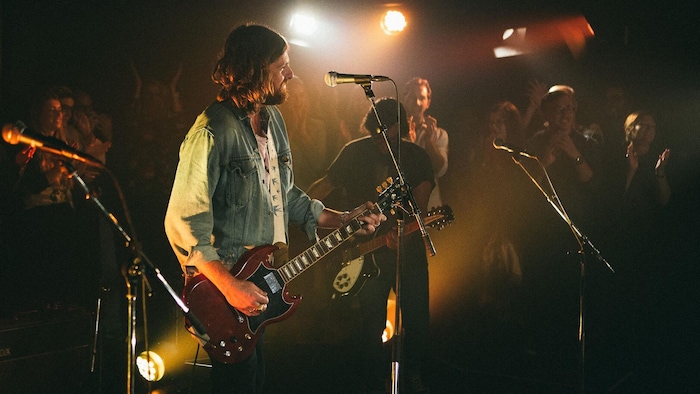 Un musicien jouant de la guitare électrique et les autres membres de son groupe jouent dans un studio, sans estrade, et il est entouré du public.