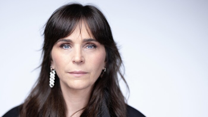 Une femme brune aux yeux bleus regarde droit devant elle.