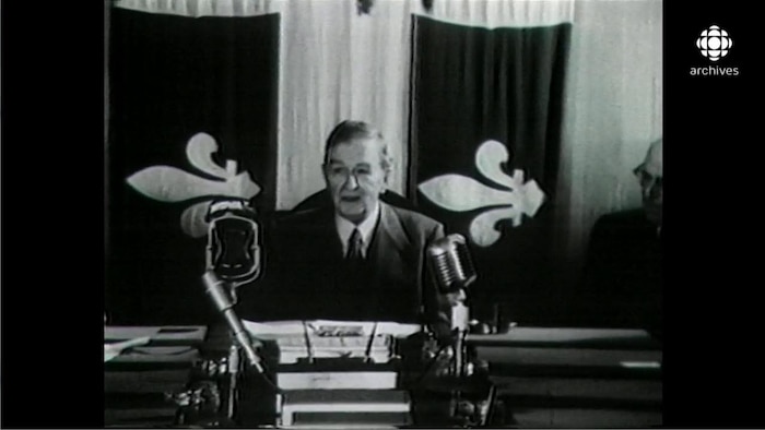 Image en noir et blanc de Maurice Duplessis, assis à un bureau massif équipé de micros, avec en arrière-plan un grand drapeau du Québec.