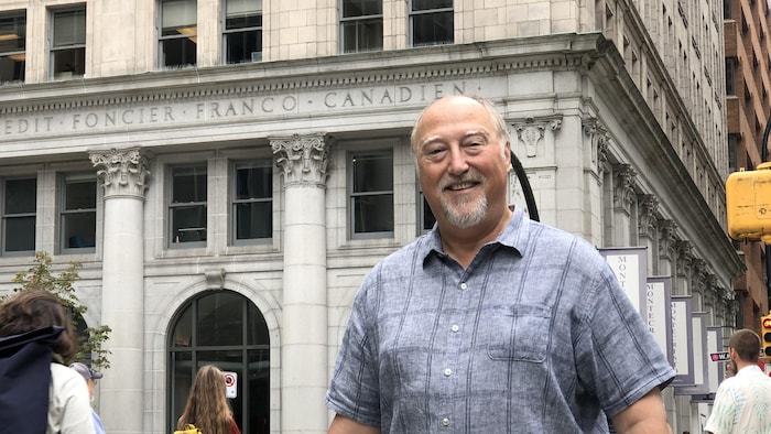 Un homme, portant une chemise à manches courtes, sourit devant la façade d'un immeuble en pierre où il est écrit : crédit foncier franco-canadien. 