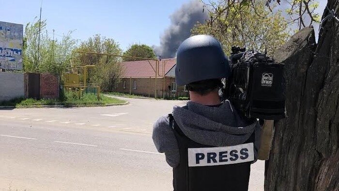 Caméra à l'épaule, il filme un bâtiment d'où s'échappe une épaisse fumée.