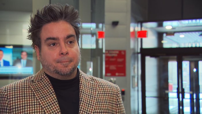 Portrait en plan moyen de Maxime Lapointe, un homme aux cheveux bruns coiffés vers le haut et portant une veste à carreaux. Il se tient dans un hall moderne avec des portes vitrées et des écrans de télévision en arrière-plan. Son expression est sérieuse et attentive.