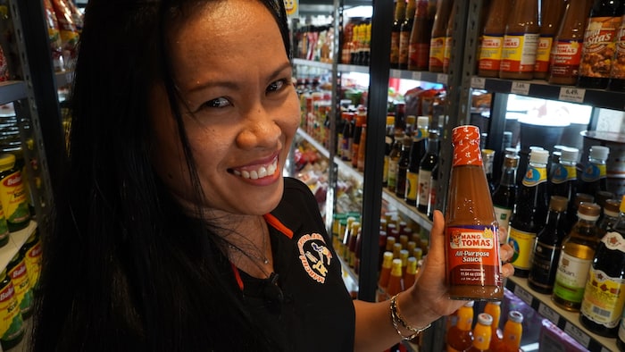 Une femme habillée en noir tient une bouteille de sauce rouge dans sa main. Elle se trouve dans une allée d'épicerie.