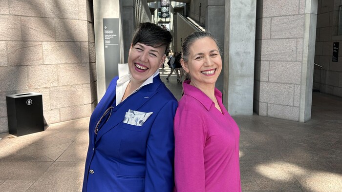 Deux femmes sont dos à dos et sourient à la caméra. L'une (à gauche) porte un wont bleu, et l'autre (à droite) porte une blouse rose.