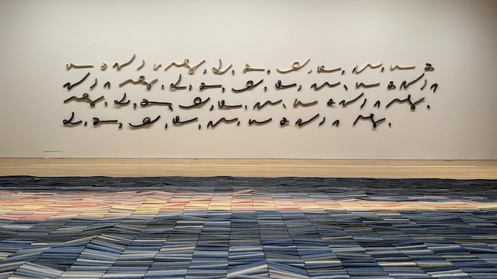 Un texte en sténographie est affiché sur le mur. En avant-plan, une «tapisserie» en perles de céramique est déposée sur un présentoir.