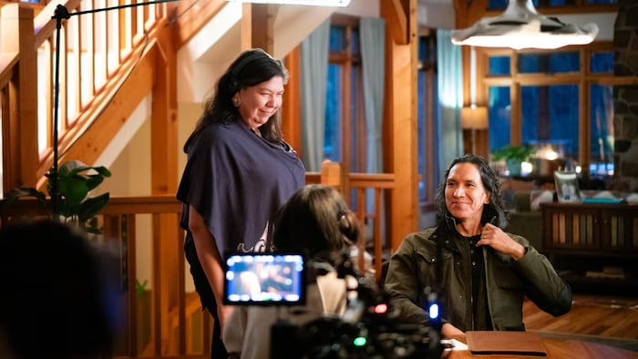 Tasha Hubbard est debout au côté de Michael Greyeyes qui est assis sur une chaise, look   à une table.
