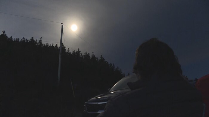 La silhouette d'une personne de dos assise dans la nature, sous un ciel sombre, la tête levée vers le soleil pendant une éclipse.