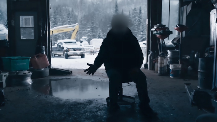 Un individu est assis sur une chaise dans un garage. Son visage est flouté. Par la porte du garage, dehors, on voit des camions sous la neige.