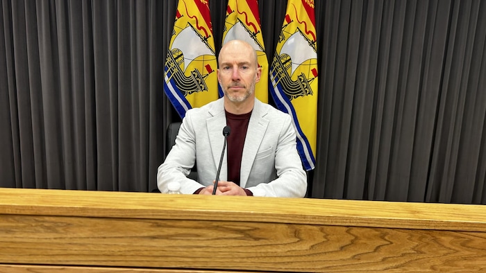 Un homme assis devant un micro, qui regarde la caméra, des drapeaux du Nouveau-Brunswick derrière lui.