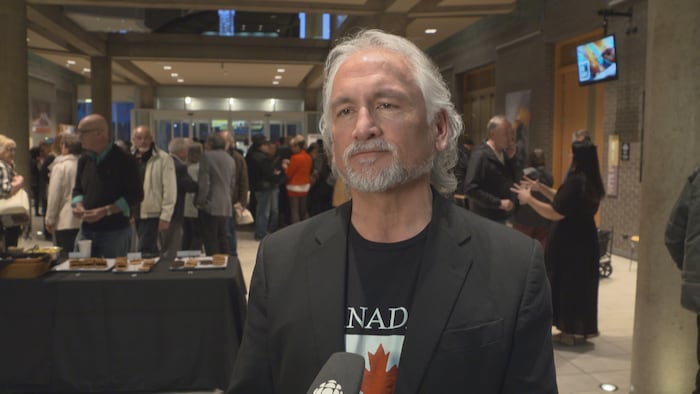 Barry Morishita se trouve dans une salle avec des dizaines de personnes derrière lui.