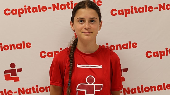 Elle pose avec son t-shirt des Jeux du Québec devant un panneau où les mots Capitale-Nationale sont inscrits plusieurs fois.