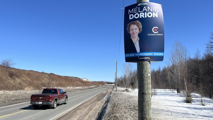 Une pancarte électorale fixée sur un poteau au bord d'une route où circule une camionnette