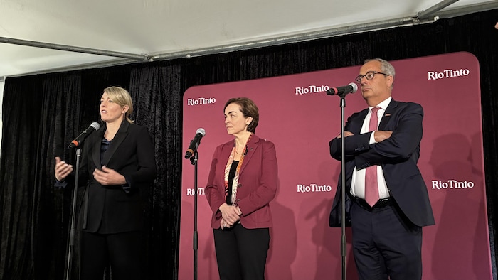 Trois personnes, deux femmes et un homme en tenue formelle, se tiennent sur une scène devant un fond rouge marqué du logo Rio Tinto. La femme de gauche s'exprime au microphone pendant que ses collègues écoutent attentivement.
