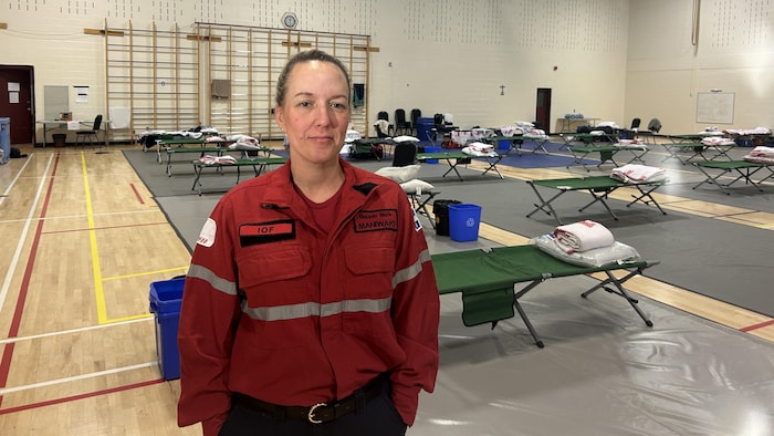 Une femme en uniforme sourit à la caméra, dans un gymnase d'école, des dizaines de lits de camp derrière elle.
