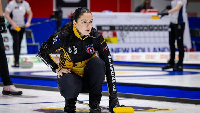Une skip en uniforme noir et jaune agenouillée sur la glace, une main au sol et l'autre sur son genou, regardant au loin.