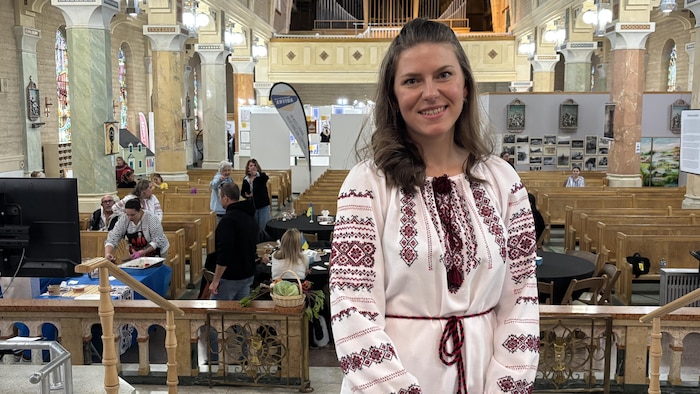 Une femme souriante, vêtue d'une blouse traditionnelle blanche à broderies rouges, pose au premier plan à l'intérieur d'une grande église.