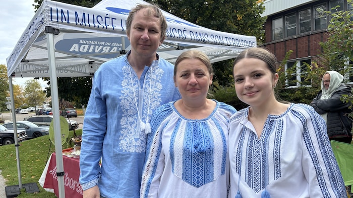 Trois personnes posent ensemble en extérieur. L'homme et les deux femmes portent des chemises traditionnelles brodées bleues et blanches.