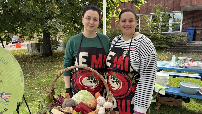 Deux femmes portant des tabliers posent derrière une table sur laquelle est posé un panier de légumes. Elles sont dehors.