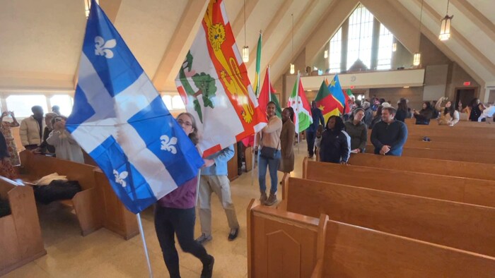 Des gens marchent à la file dans l'allée de l'église entre les bancs, en tenant des drapeaux du Québec, de l'Île-du-Prince-Édouard, et de pays d'Afrique, comme le Cameroun, le Burundi et la République démocratique du Congo.