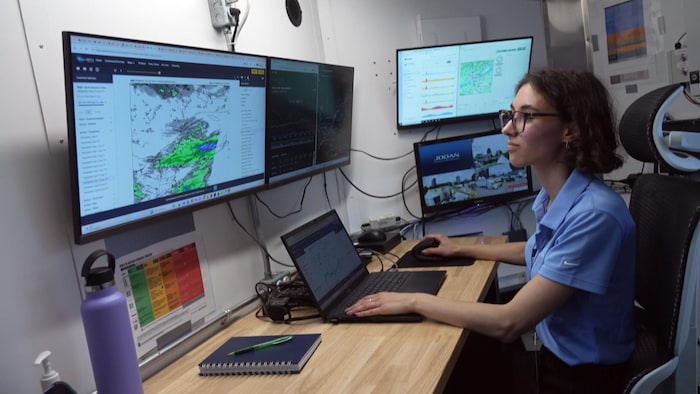 Une femme est assise à un bureau où elle regarde des écrans. 