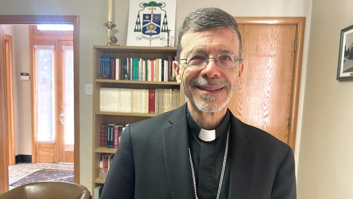 Un évêque en vêtements sacerdotaux noirs, portant un col romain blanc, photographié debout dans son bureau.