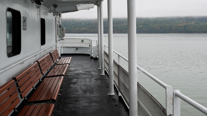 Un pont de bateau vide avec des bancs en bois longe l'eau face à une rive verdoyante sous un ciel couvert.