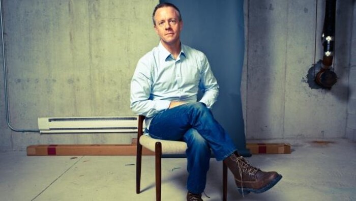 Un portait de Michael Bryant assis sur une chaise. Il a une chemise bleue et des jeans et les jambes croisées. 