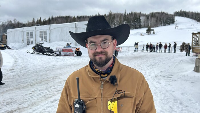 Un homme coiffé d'un chapeau de cowboy est debout dehors en hiver. Il a un walkie-talkie et des lunettes de ski accrochées aux poches avant de sa veste.