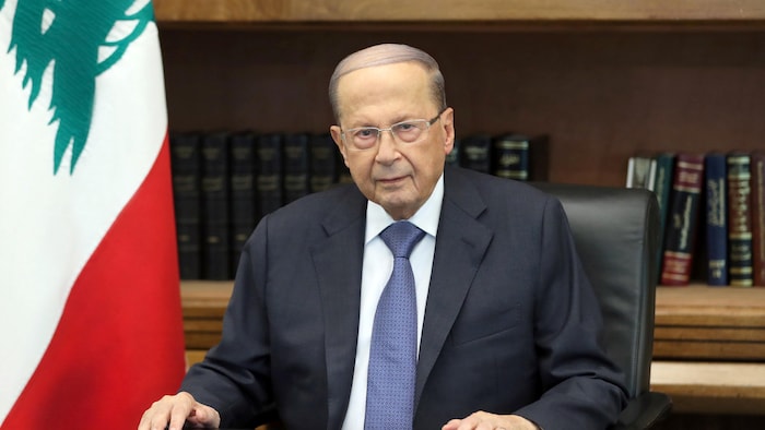 Michel Aoun est assis derrière une table, devant une bibliothèque, à côté d'un drapeau du Liban. 