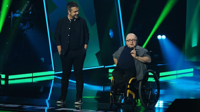 Un homme debout et un autre en fauteuil roulant sur une scène.
