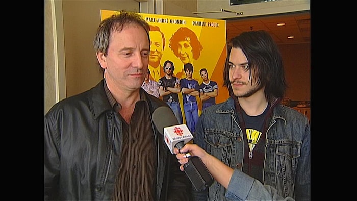 Michel Côté au micro de Radio-Canada aux côtés de Marc-André Grondin devant une affiche du film.